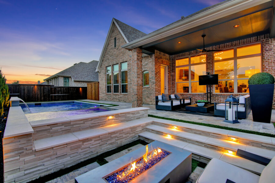 This lifestyle image captures the essence of luxury living by showcasing a beautiful outdoor space at sunset. A modern fireplace takes center stage, surrounded by comfortable chairs for relaxation and conversation. In the background, a covered patio with additional seating provides the perfect spot for enjoying a drink or meal. The blue pool sparkles with pool lighting, inviting you for a refreshing swim on a warm evening. The tranquil setting and modern design make this luxurious home an ideal retreat for entertaining or relaxation.